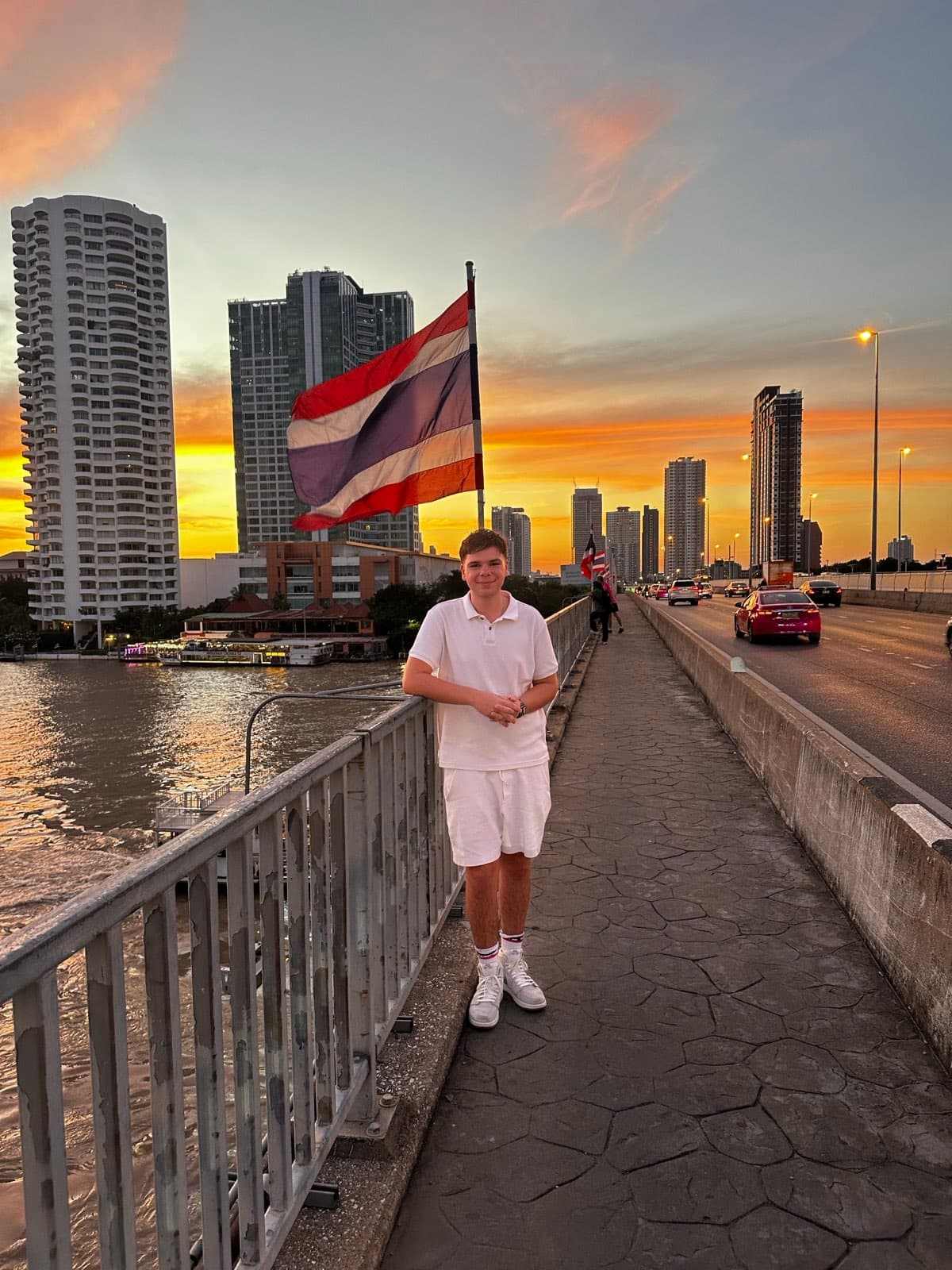 Standing on a bridge walkway above a city river at sunset, with a flag flying and high-rise towers on the far bank.